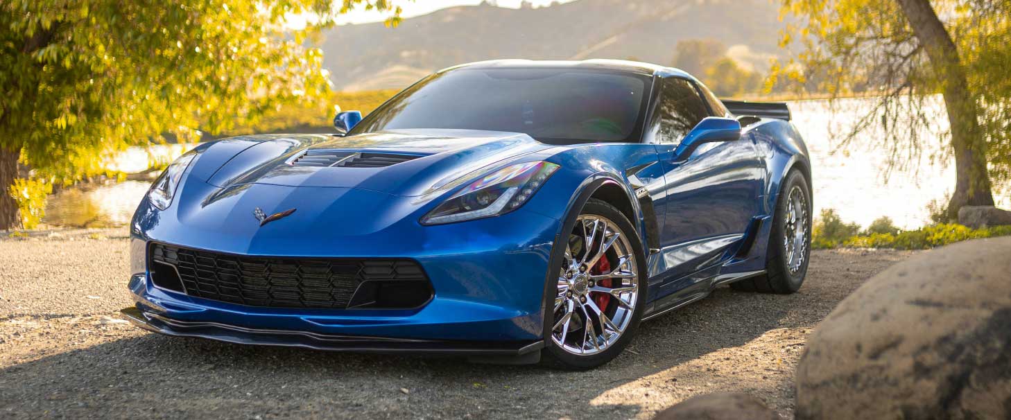 An image of a blue sports car parked at the edge of a lake, framed by trees with green foliage